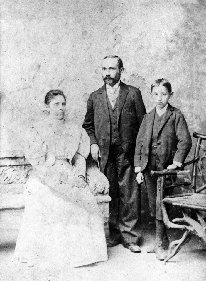 Czarno białe zdjęcie przedstawiające rodzinę: siedząca kobieta w białej sukni, stojący mężczyzna z brodą i stojący mały chłopiec.