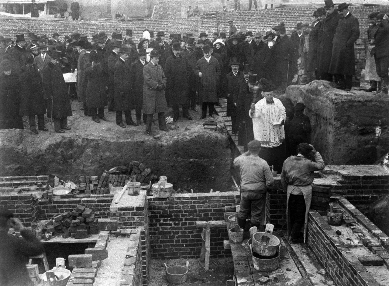 Czarno białe zdjęcie z grupą ludzi na terenie budowy, z księdzem pomiędzy nimi.