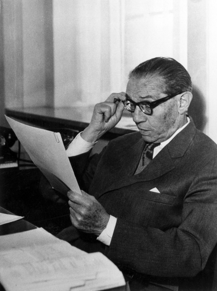 Czarno białe zdjęcie starszego mężczyzny w okularach siedzącego przy biurku trzymającego kartkę papieru.