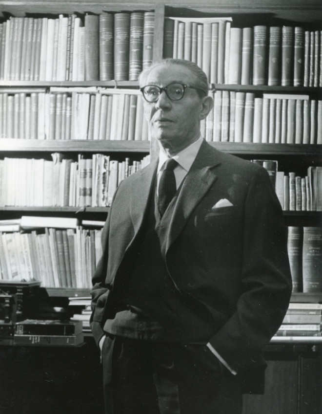 Czarno białe zdjęcie  stojącego na tle biblioteczki mężczyzny w okularach.