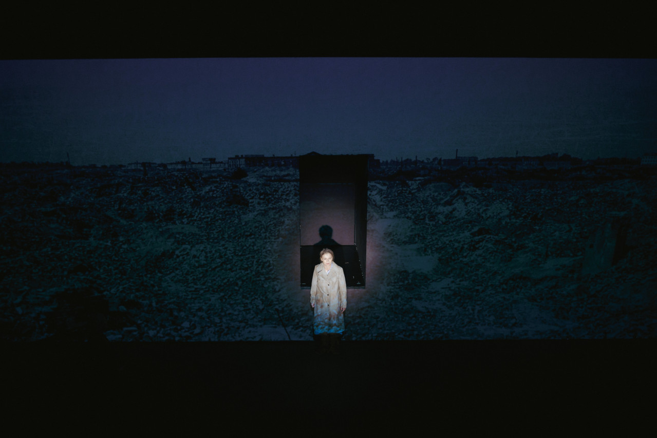 Młoda kobieta w zniszczonym płaszczu siedzi na progu otworu w ścianie. Za nią ciemność. Na ścianie projekcja - miasto w ruinie. 