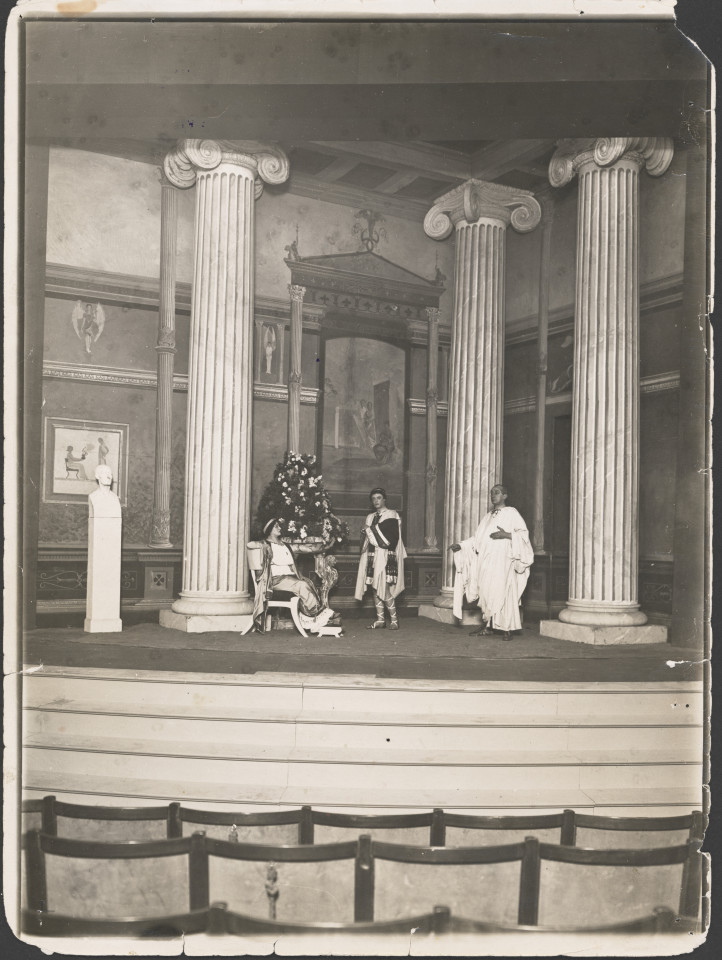 Na czarno-białym zdjęciu grupa aktorów na scenie pośród scenografii teatralnej spektaklu Irydion.