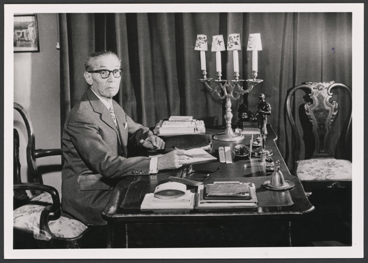 Na czarno-białym zdjęciu Arnold Szyfman w swoim gabinecie w domu przy ulicy Prezydenckiej, około roku 1957. Starszy mężczyzna w okularach przy biurku ze świecznikiem.