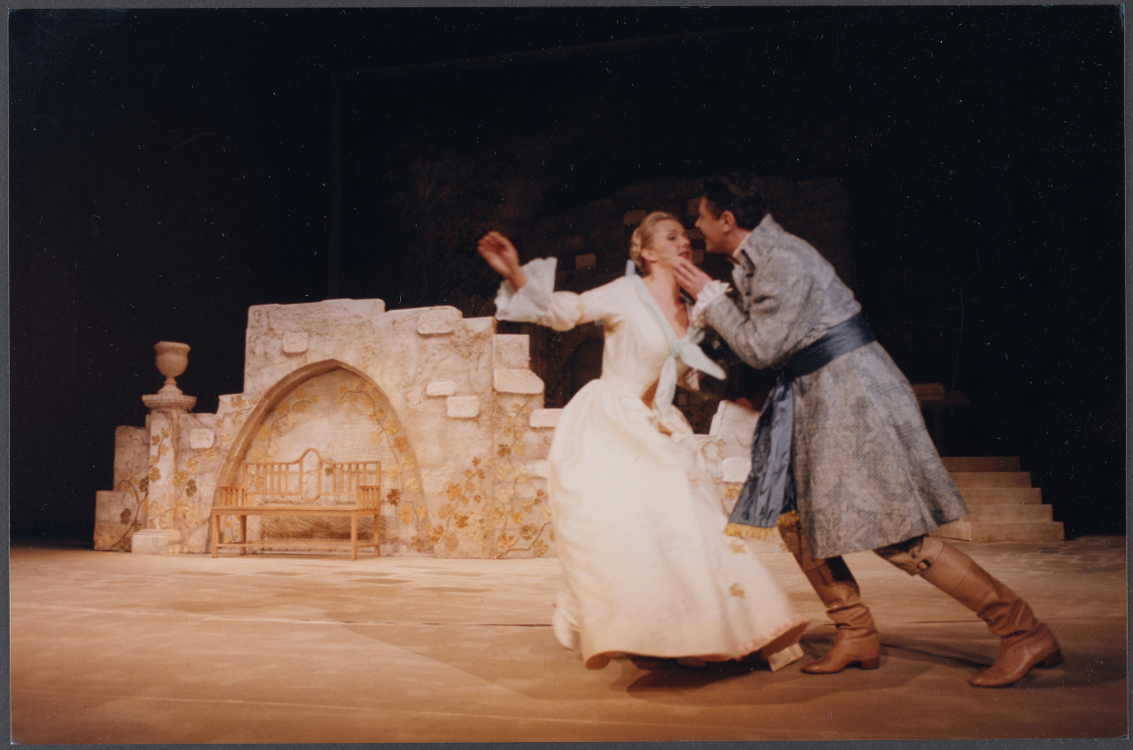 Kobieta w białej sukni i mężczyzna w niebieskim surducie na tle muru i ławeczki, scenografii teatralnej.