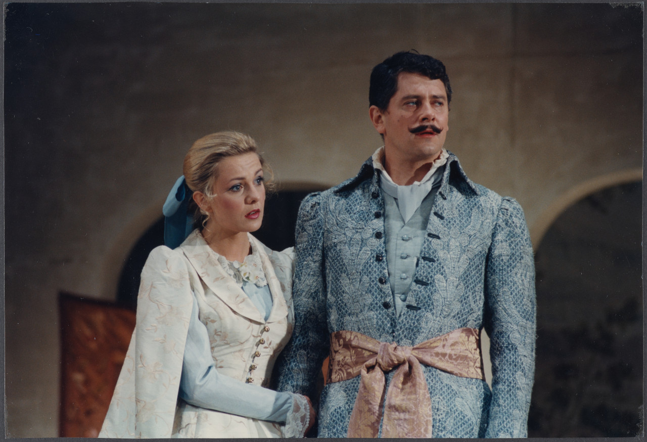 Kobieta w białej sukni i mężczyzna w niebieskim surducie na tle muru, scenografii teatralnej.