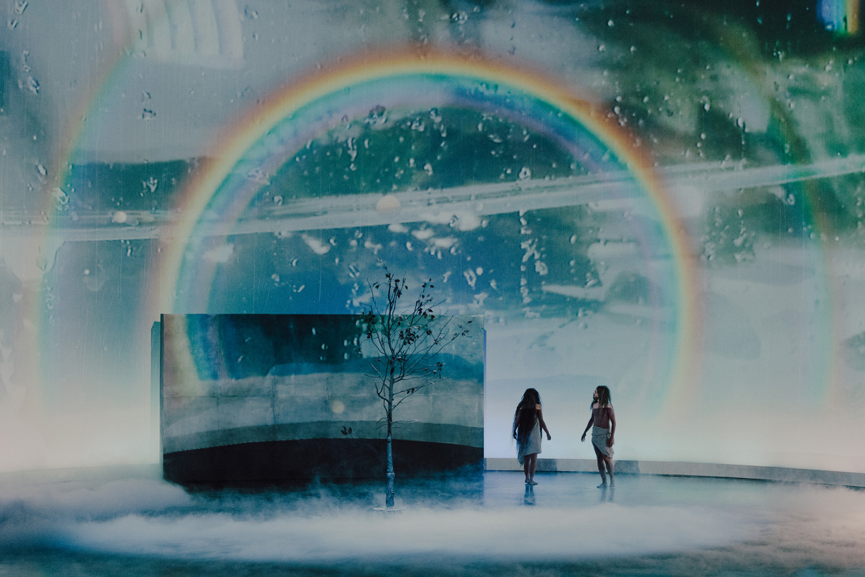 Scena ze spektaklu „Rękopis znaleziony w Saragosie. Dziedzictwo Gomelezów”: dwie postacie na spowitej mgłą scenie, pod świetlistą tęczą, obok samotnej rośliny.