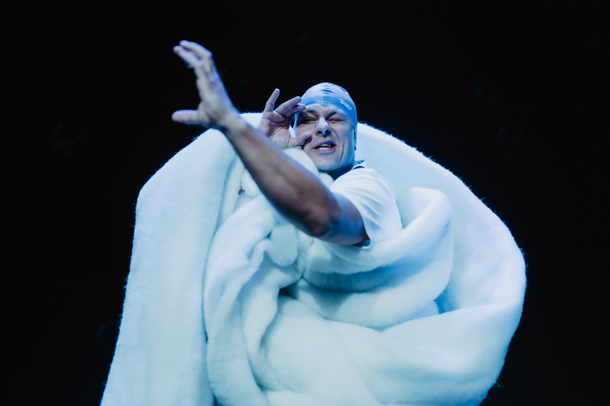 Postać owinięta w miękki, biały kokon wyłania się z mroku sceny, gestem dłoni jakby zapraszając widza do wejścia w świat dziecięcej wyobraźni i snu.